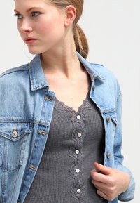 Denim jack over een grijze mouwloze top met kantafwerking en knoopdetails. De jas heeft een klassieke snit en metalen hardwareaccenten.