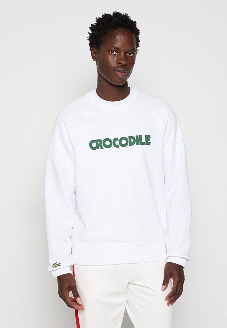 Weißes Sweatshirt mit grüner "CROCODILE"-Schrift, Rundhalsausschnitt, Raglanärmeln und kleinem grünen Logo am Saum. Glatte Stoffoberfläche.