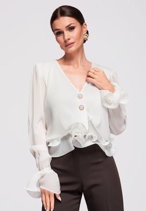 Femme aux cheveux tirés en arrière portant un chemisier blanc plissé avec des poignets et un ourlet à volants, des boutons dorés, un pantalon noir et de grandes boucles d'oreilles dorées.