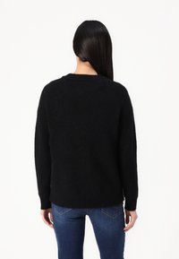Frau mit langen, glatten schwarzen Haaren, die einen schwarzen Pullover und blaue Jeans trägt und mit dem Rücken zu einem schlichten weißen Hintergrund steht.