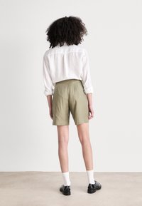 Pantalones cortos verde oliva con un corte entallado, que presentan una textura suave y pliegues en la parte trasera. Combinados con una camisa de lino blanca y zapatos negros.