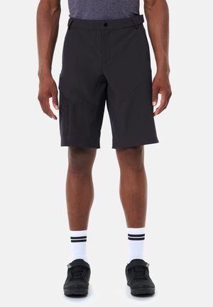 Mann steht, trägt schwarze knielange Shorts, graues Hemd, weiße Socken mit schwarzen Streifen und schwarze Sportschuhe vor einfachem Hintergrund.