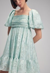 Valkoinen, polvipituinen mekko, joka on valmistettu teksturoidsesta kankaasta, puffihihnoilla ja neliönmuotoisella kaula-aukolla, jossa on kerrokselliset röyhelöt helmassa.