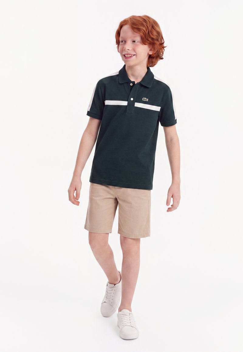 Garçon roux marchant, portant un polo foncé à rayures blanches, un short beige et des baskets blanches, souriant et regardant vers la droite.