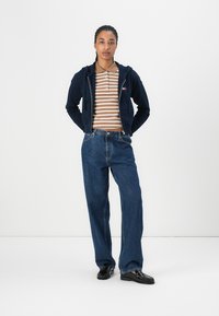 Felpa con zip blu sopra una polo a righe marroni e bianche, abbinata a jeans blu larghi e scarpe nere. Un outfit semplice e casual.