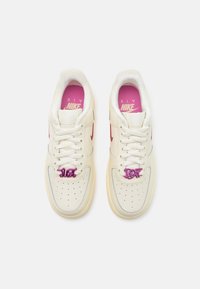 Baskets Nike Air blanches avec semelles intérieures roses, logos swoosh rouges et breloques métalliques violettes "Just Do It" sur les lacets, vues de dessus.