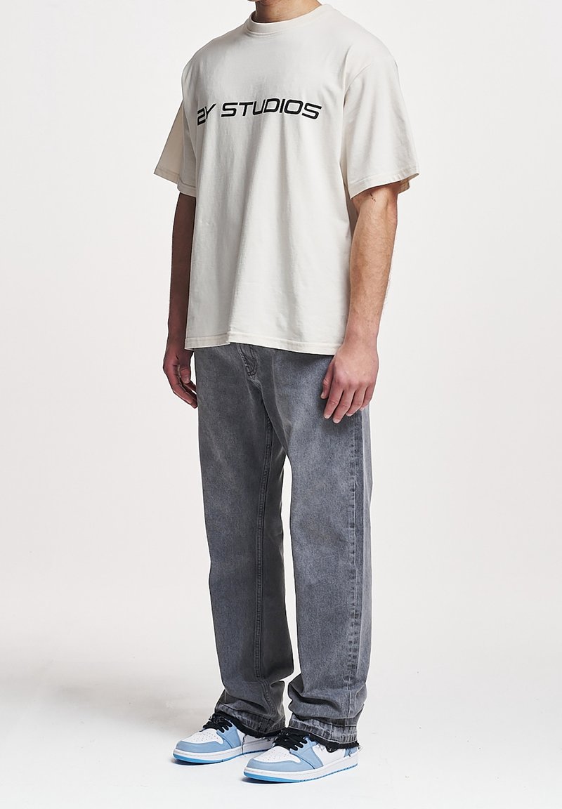 Homme portant un t-shirt blanc oversize avec le texte "2Y STUDIOS", jean gris et baskets blanches, bleues et noires, debout devant un fond uni.