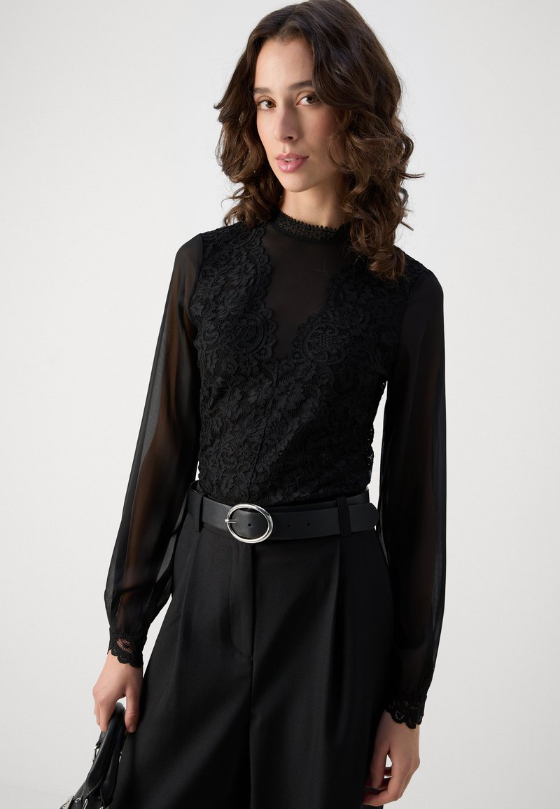 Top de encaje negro con mangas largas transparentes y cuello alto. Combinado con pantalones negros de cintura alta y un cinturón negro ancho con hebilla plateada.