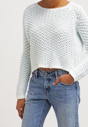 Kobieta ubrana w jasnoniebieski, krótki sweter dziergany oraz wysokiej talii niebieskie dżinsy, z jedną ręką w kieszeni na białym tle.