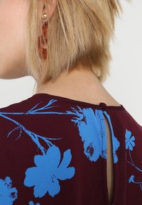 Robe fleurie en tissu bordeaux avec des motifs de fleurs bleues, dos avec une ouverture en forme de goutte et fermeture par bouton au niveau du cou. Finition texturée.