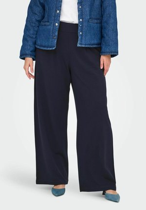 Persoon in een wijde zwarte broek, blauwe gewatteerde jas, witte shirt en blauwe puntige hakken, staand tegen een witte achtergrond.