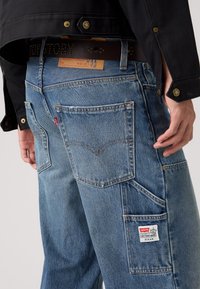 Pantalones cargo de mezclilla azul claro con múltiples bolsillos, detalles de costura y una etiqueta de marca. Se aprecian acentos de hardware revestido.