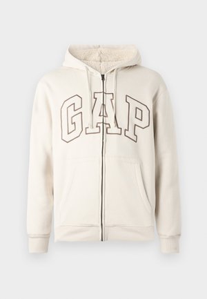 Beige zip-up hoodie with fleece-lined hood, drawstrings, front pockets, and large outlined "GAP" letters across chest.