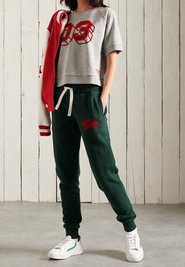 Grijze cropped sweatshirt met rode grafische "03," gecombineerd met donkergroene joggers. Witte sneakers hebben groene accenten. Achtergrond is van hout.