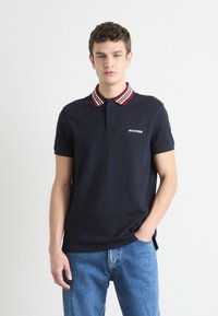 Σακάκι ναυτικού χρώματος με γιακά ριγέ σε κόκκινο, άσπρο και μπλε, που διαθέτει κλείσιμο με κουμπιά στο λαιμό και λογότυπο στο αριστερό στήθος. Συνδυάζεται με μπλε τζιν.