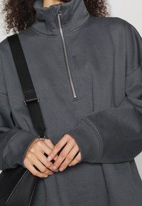 Pull-over gris charbon avec col montant, fermeture éclair et manches amples. Sangle noire ajustable visible, mains jointes devant.