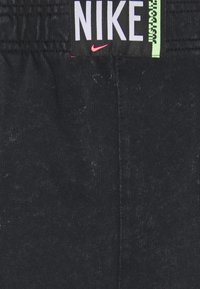Juodas „Nike“ juosmuo su storu elastingu juostu. „NIKE“ išryškintai nurodyta baltu šriftu, o „JUST DO IT“ žaliu, taip pat yra mažas rožinis logotipas.
