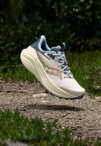 Beige ja harmaa juoksukenkä, jossa on teksturoitu päällinen, heijastavat yksityiskohdat ja paksu, pehmustettu pohja. Siinä on nauhallinen sulkeminen ja tukeva muotoilu.