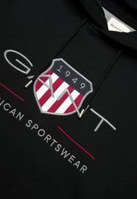 Felpa con cappuccio nera caratterizzata da un logo scudetto grigio e rosso con la scritta "GANT" e "1949", oltre a una linea orizzontale rossa sotto "AMERICAN SPORTSWEAR."