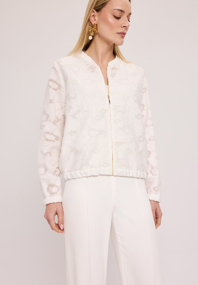 Biała bomberka z teksturowanym wzorem, z zamkiem błyskawicznym i marszczonym dołem. Długie, przezroczyste rękawy oraz elastyczne mankiety dodają struktury i detali.
