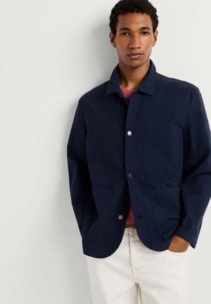 Junger Mann mit lockigem Haar, der eine marineblaue, geknöpfte Jacke über einem roten Hemd und weißen Hosen trägt und vor einem einfarbigen hellen Hintergrund steht.