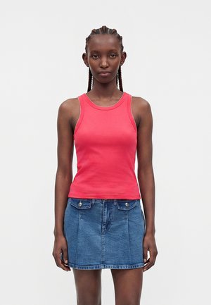 Frau mit geflochtenem Haar, die ein pinkfarbenes ärmelloses Oberteil und einen blauen Jeansrock trägt, steht vor einem schlichten weißen Hintergrund.