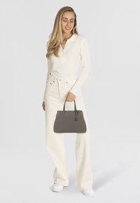 Borsa grigia con doppi manici e sottili dettagli metallici. Cuoio liscio, design strutturato, tenuta da una persona in abbigliamento bianco e pantaloni larghi.