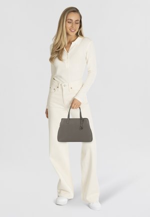 Sac à main gris avec deux poignées et des accents de quincaillerie discrets. Cuir lisse, design structuré, tenu par une personne en tenue blanche et en pantalon ample.