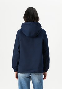 Marineblå hettejakke med elastiske mansjetter og en avslappet passform, laget av glatt stoff, vist bakfra med blå denimjeans.