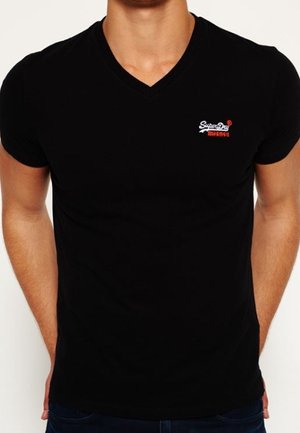 Mężczyzna noszący czarną koszulkę z dekoltem w serek z małym białym i czerwonym logo SuperDry na lewej piersi.