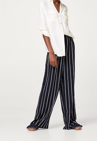 Chemise blanche à boutons, avec les manches retroussées, associée à un pantalon large à rayures verticales bleu marine. Des sandales avec des accents froncés complètent la tenue.