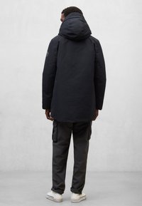 Ciemna kurtka z kapturem o teksturowanym wykończeniu, długim kroju i widocznym przeszyciem, zestawiona z szarymi spodniami cargo i białymi tenisówkami.