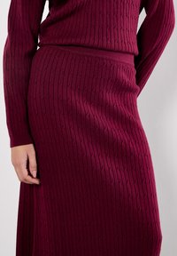Burgundy rippad stickad klänning med figurnära midja och långa ärmar. Texturerat mönster med en mjuk yta som framhäver silhuetten.