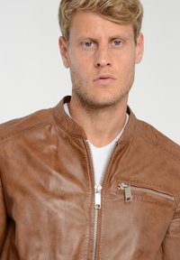 Bruine leren jas met zilveren ritssluiting en zakdetails. Heeft een ronde halslijn en een zachte textuur, gedragen over een wit T-shirt.