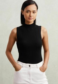 Melna bezpiedurkņu blūze ar augstu kakla izgriezumu, ribota tekstūra un vertikālām šuvēm, apvienota ar baltām augstām biksēm.