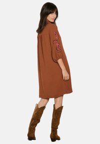 Robe marron surdimensionnée avec des manches bouffantes et broderie florale rose sur une manche, associée à des bottes en suede marron jusqu'aux genoux.