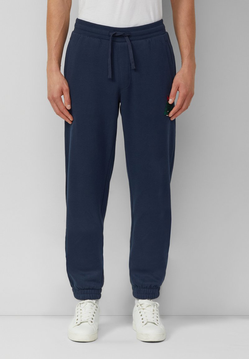 Mężczyzna ubrany w granatowe spodnie dresowe ze sznurkiem i białe trampki, stojący na tle jednolitego jasnoszarego tła.