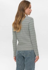 Ženska z dolgimi valovitimi lasmi, oblečena v dolge rokave črno-belo progo in modre jeans, stoji bočno.