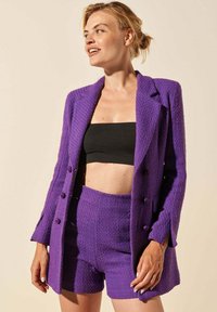Blazer violet texturé avec des shorts taille haute assortis. Comprend des fermetures à bouton et une coupe ajustée. Porté sur un crop top noir.