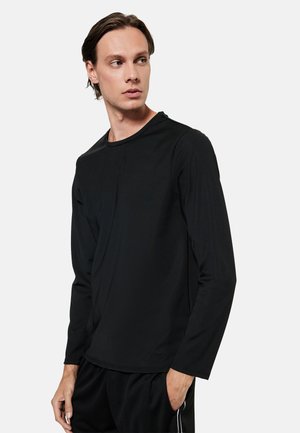 Man met een lichte huid en donker haar kijkt naar links, draagt een effen zwart shirt met lange mouwen en een zwarte broek met witte strepen, hand in de zak.