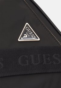 Svart nylonaccessoar med en triangulär silverlogotyp med texten "GUESS U.S.A.". Inkluderar en texturerad svart märkessnodd.