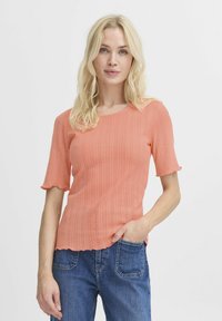 ferskenfarvet ribstrikket top med korte ærmer, bølgede kanter og rund halsudskæring. Bæres med blå denim jeans, der fremhæver en tætsiddende pasform.