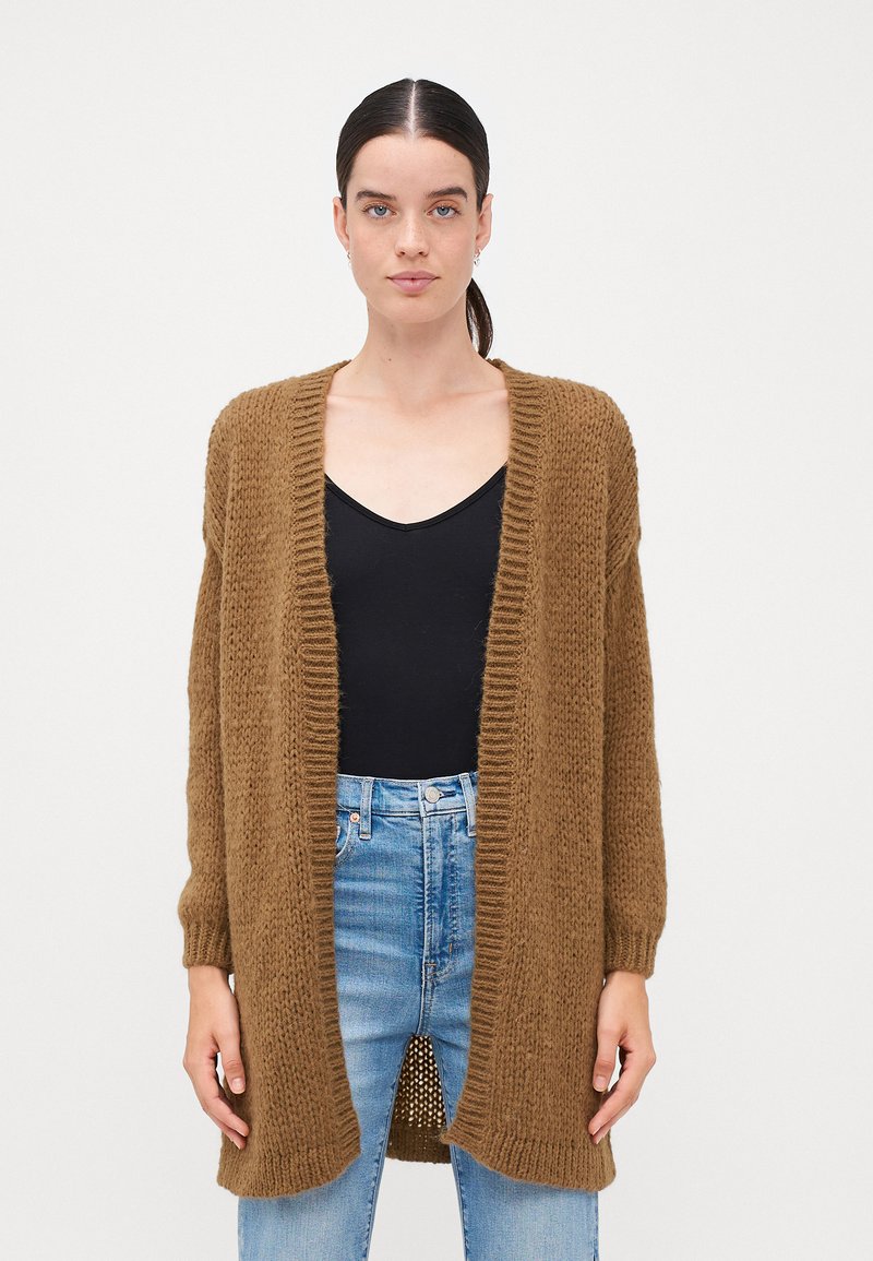 Cárdigan marrón de punto con un ajuste holgado, frontal abierto, bordes acanalados y mangas largas, combinado con una blusa negra y jeans azules.