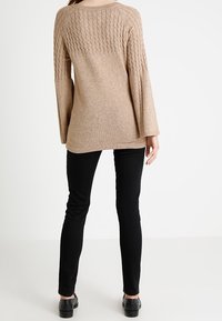 Beżowy sweter dzianinowy z teksturowanym wzorem na plecach, z długimi dzwonami rękawów, zestawiony z czarnymi dopasowanymi spodniami i czarnym obuwiem.