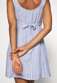 Robe rayée avec des lignes verticales bleues et blanches, un dos échancré et une ceinture nouée à la taille. Tissu doux avec une coupe décontractée.