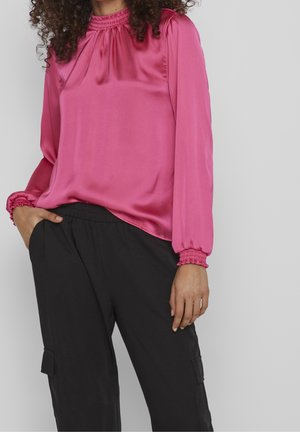 Kobieta nosząca różową satynową bluzkę z długim rękawem, marszczonym kołnierzem i mankietami, zestawioną z luźnymi czarnymi spodniami typu cargo, ręka w kieszeni.