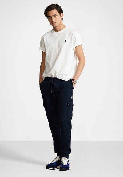T-shirt bianco in cotone con maniche arrotolate, pantaloni cargo blu in felpa con tasche e scarpe da ginnastica blu con accenti bianchi.