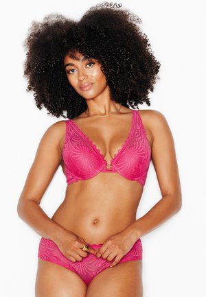 Donna con i capelli ricci che indossa un reggiseno in pizzo rosa brillante e biancheria intima abbinata, in piedi su sfondo bianco.