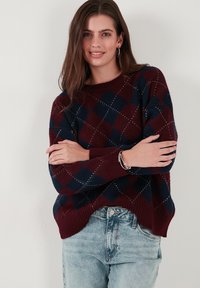 Pull en tricot à motif argyle bourgogne et bleu marine avec des poignets et un ourlet côtelés. Associé à un jean bleu clair, présentant une coupe décontractée.