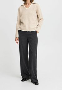 Pull beige avec col et ourlet festonnés, associé à un pantalon large gris foncé en tissu texturé et chaussures en cuir verni noir.